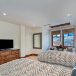 Bedroom with twin beds, chevron-patterned bedding, wooden dresser with a TV, a wall lion mask, large windows with mountain views, and cozy seating.
