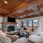 Luxury villa living room with wooden ceiling, stone fireplace, plush sofas, and large windows overlooking snowy mountains.