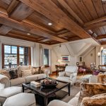 Luxury living room with wooden ceiling and large windows overlooking snowy mountains, featuring plush sofas, a coffee table, and a dining area with a chandelier.
