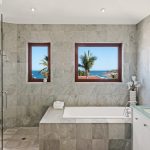 Luxurious bathroom with stone tile design, featuring a glass shower, bathtub, and ocean view through two windows.