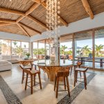 Luxury villa interior with a rustic wooden ceiling, elegant furniture, and large glass windows overlooking a tropical landscape.