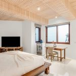 Bedroom with a modern wood ceiling, bed, TV, wooden desk, and windows overlooking a patio.