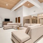 Spacious living area with beige sectional sofas and wooden bunk beds, featuring a sleek stainless steel refrigerator and a TV on a dresser, under a wooden ceiling.