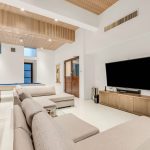 Spacious modern living room with beige sectional sofa, large flat-screen TV, bunk beds, and a pool table, featuring wood panel ceiling and minimalistic decor.