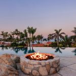 A luxurious swimming pool with a fire pit, surrounded by palm trees, overlooking the ocean at sunset.
