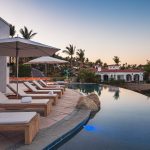 Outdoor pool area with lounge chairs and umbrellas at a luxury villa in Los Cabos, with palm trees and villas in the background.
