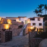 A luxurious villa with stone walls and palm trees, illuminated at dusk.