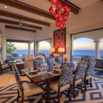 Elegant dining and living area in a luxury villa with ocean views, featuring a decorative chandelier, blue-patterned chairs, and large windows overlooking a sunset.