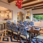 Elegant dining and living area in a luxury villa with blue and white decor, featuring a large dining table, patterned chairs, open kitchen with bar seating, and an adjoining outdoor patio.