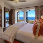 Luxurious bedroom with ocean view, decorated with orange and white pillows, wooden furniture, and large windows overlooking the beach at a villa in Los Cabos, Mexico.