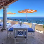 Outdoor terrace with white furniture, blue cushions, and striped umbrellas overlooking the ocean.