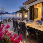 Outdoor dining area with a colorful table setting, surrounded by chairs and flowers, overlooking the ocean with lounge chairs and umbrellas.