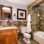 Luxury bathroom with natural stone tile, wooden vanity, framed mirror, glass-enclosed bathtub, and decorative wall art.