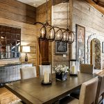 Dining room with wooden walls and a rustic chandelier, featuring a modern table with beige chairs and decorative candles.