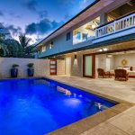 Modern house with an illuminated pool, patio seating, and an open bedroom door at dusk.