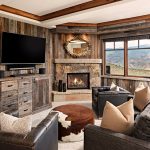 Luxury villa living room with rustic wooden walls, a stone fireplace, large TV, cozy seating, and mountain views through the window.