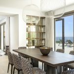 Modern dining room with patterned chairs, a wooden table, large pendant lights, and glass doors leading to a seaside balcony.