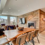 Interior of a luxury villa with modern decor, featuring a wooden dining table, leather chairs, a fireplace, and large windows overlooking snowy mountains.
