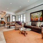 Luxurious living area with modern decor, featuring a leather sofa, armchair, glass coffee table, dining table, bar stools, and a vibrant painting, set in a bright, open-concept space.