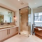 Luxury bathroom with marble bathtub, glass shower, and large vanity with dual sinks, overlooking snowy mountain landscape through large windows.