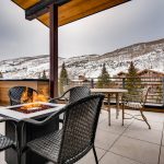 Outdoor patio with a fire pit, surrounded by wicker chairs and a table, overlooking snow-covered mountains and trees in Vail, Colorado.
