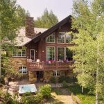 A luxurious villa with large windows and a stone chimney, surrounded by trees, featuring a deck with flowers and a hot tub on the patio.