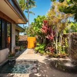 Tropical outdoor area of a luxury villa with a small pool, lush plants, and a stone pathway leading to a wooden gate.