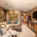 Luxurious living room with beige sofas, decorative pillows, and tropical-themed artwork on the walls, featuring a large flat-screen TV and elegant wooden furniture.