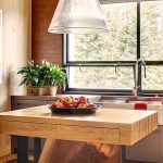 Modern kitchen interior with a wooden island, a bowl of apples, a large window with a wooded view, and a Campbell's tomato soup poster on the wall.