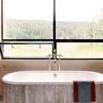 A luxurious bathroom with a freestanding bathtub, large window with a scenic view, and wooden cabinetry.