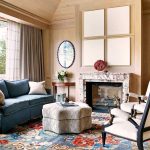 Luxurious living room with a blue sofa, patterned ottoman, plush chairs, and a marble fireplace, adorned with stylish decor and a colorful rug. Large windows allow natural light and a view of greenery.