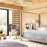 Luxurious bedroom interior with a four-poster bed, patterned textiles, woven chair, and large windows overlooking a wooded area.