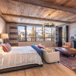 Luxurious bedroom in a villa with a large window view of snowy mountains, featuring a cozy fireplace, leather chairs, and rustic wooden interior.