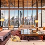 Luxurious living room with large windows showcasing a snowy mountain view, featuring elegant sofas, armchairs, and a coffee table in a wooden and stone interior.