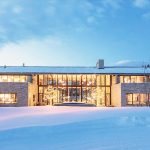 Luxurious villa with large windows illuminated at night, surrounded by snow-covered landscape and trees.