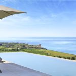 Infinity pool overlooking a coastal landscape with lush greenery, a golf course, and a tower near the ocean under a clear blue sky, partially shaded by an umbrella.