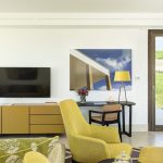 Modern living room with a mustard yellow theme, featuring a TV mounted on the wall, artistic wall decor, a yellow armchair, a console, and a side table with a lamp. There is a door with a view of greenery outside.