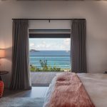 Luxury villa bedroom with ocean view, featuring a modern chair, lamp, and large windows overlooking the Caribbean sea.