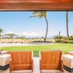 Luxury villa patio with wooden chairs overlooking a lush lawn, pool, and a scenic beach view with palm trees in Hawaii.