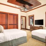A luxurious bedroom with two beds, wooden accents, a ceiling fan, and a flat-screen TV, viewed through large windows showing tropical plants.