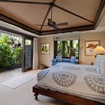 Luxurious bedroom with elegant decor, featuring a king-sized bed, ceiling fan, large windows, and sliding doors opening to lush tropical gardens.