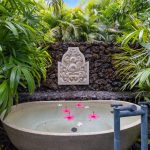 An outdoor stone bathtub filled with water and floating red flowers, surrounded by lush green tropical plants and a decorative stone relief on a rock wall.