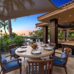 Outdoor dining area of a luxury villa with wooden furniture, overlooking tropical plants and a beach sunset in Hawaii.