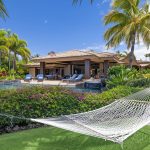 Luxury villa with a swimming pool surrounded by tropical greenery, a hammock in the foreground, and a clear blue sky.