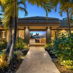 Elegant luxury villa entrance surrounded by lush greenery, opening to a beachfront view with palm trees at sunset.