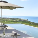 Infinity pool overlooking a coastal landscape with lounge chairs and an umbrella.