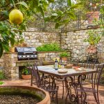 Outdoor stone patio with lemon trees, a dining table set with wine and food, and a barbecue grill in the background.