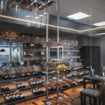 Glass-enclosed wine room with racks of wine bottles and wine glasses, overlooking a scenic ocean view.
