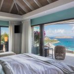 Bedroom with ocean view in a luxury villa, featuring a bed, seating area, and large glass doors opening to a balcony overlooking a beach and island.