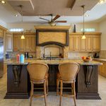 Luxurious kitchen interior with wooden cabinetry, granite countertops, two bar stools, and modern appliances.